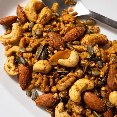 Golden toasted almonds, cashews, and walnuts glistening with a sweet and spicy maple glaze, scattered across a white surface with vibrant turmeric and paprika hues.