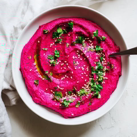 A vibrant bowl of Roasted Beet Hummus garnished with olive oil, parsley, and sesame seeds, perfect for dipping crisp vegetables.  